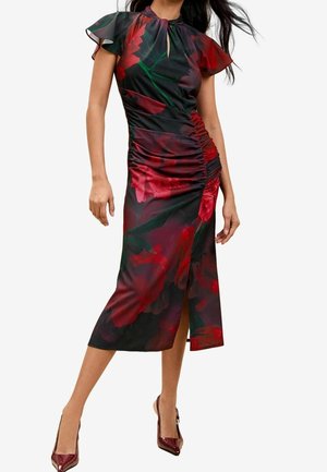 Kobieta w czarnej sukience midi z czerwonym kwiatowym wzorem, z falbanowymi rękawami, marszczeniem po bokach, przednim rozcięciem i pasującymi ciemnoczerwonymi szpilkami.