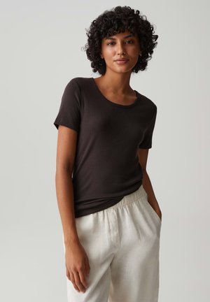 Vrouw met krullend haar die een aansluitend zwart T-shirt met korte mouwen en losse witte broek draagt tegen een effen grijze achtergrond.