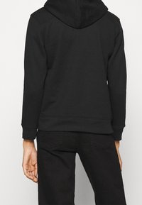 Sweat à capuche noir avec une grande capuche, des poignets côtelés et un ourlet droit, fabriqué en matériau doux. Coutures latérales visibles et sans graphismes.