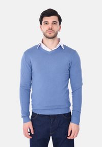 Maglione blu lavorato a maglia con scollo a V, maniche lunghe e polsini a coste. Indossato sopra una camicia bianca con colletto, abbinato a jeans in denim scuri.