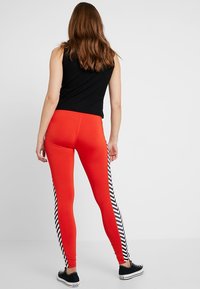 Röda leggings med ett svart och vitt zigzag-mönster på sidan, kombinerat med en svart ribbad topp och svarta sneakers.