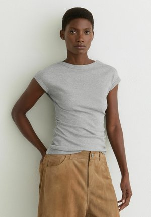 Donna con capelli corti che indossa un top grigio aderente senza maniche e pantaloni in camoscio beige a vita alta, in piedi contro un muro bianco.