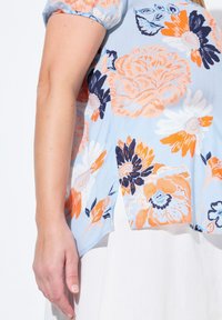 Blusa floreale azzurro chiaro con motivi di fiori arancioni e navy, maniche corte a palloncino e una vestibilità comoda con dettagli a spacco laterale.