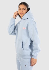 Sudadera con capucha de color azul claro, hecha de una tela suave, que presenta un bolsillo frontal y un pequeño logo rosa en el pecho. Puños y dobladillo de canalé.