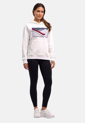 Femme debout portant un sweat à capuche blanc avec un motif de montagne et de ski, des leggings noirs et des baskets blanches sur un fond uni.