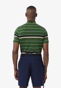 Polo verde a righe con strisce beige e nere, abbinato a pantaloni shorts blu navy. La maglietta presenta un colletto e maniche corte.