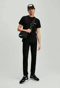 Černé tričko a kalhoty, černá crossbody taška s bílým logem, černá čepice s bílou nášivkou a lesklé černé tenisky s modrými akcenty.