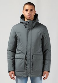 Jaunas vyras dėvi pilk�ą žieminę striukę su keliais kišenėmis ir šviesiai mėlynas džinsus, stovi prieš paprastą foną.