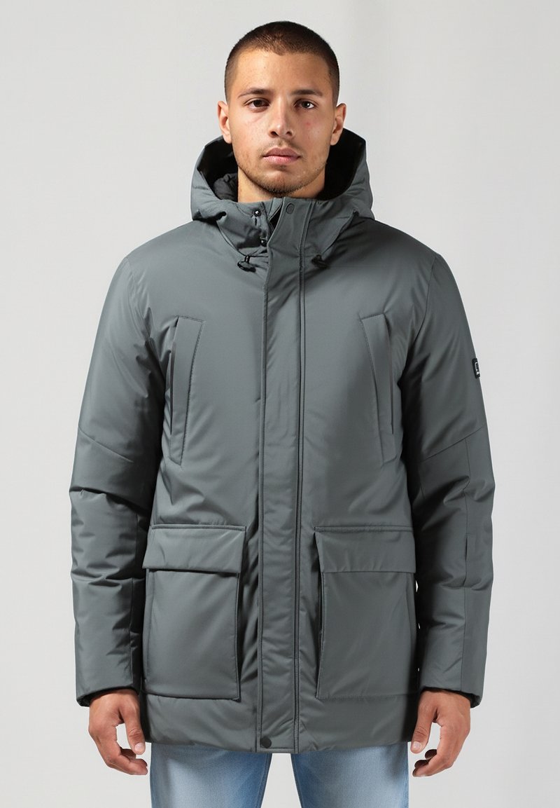 Jaunas vyras dėvi pilką žieminę striukę su keliais kišenėmis ir šviesiai mėlynas džinsus, stovi prieš paprastą foną.