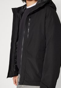 Svart puffjacka med dragkedja fram, justerbar huva och invändig foder. Har sidofickor och en bröstficka med dragkedja för förvaring.