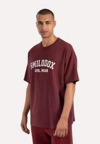 Bordeaux oversized t-shirt met de tekst "SMILODOX ATHL. WEAR" in het wit. Korte mouwen, ronde halslijn, katoenmix stof.