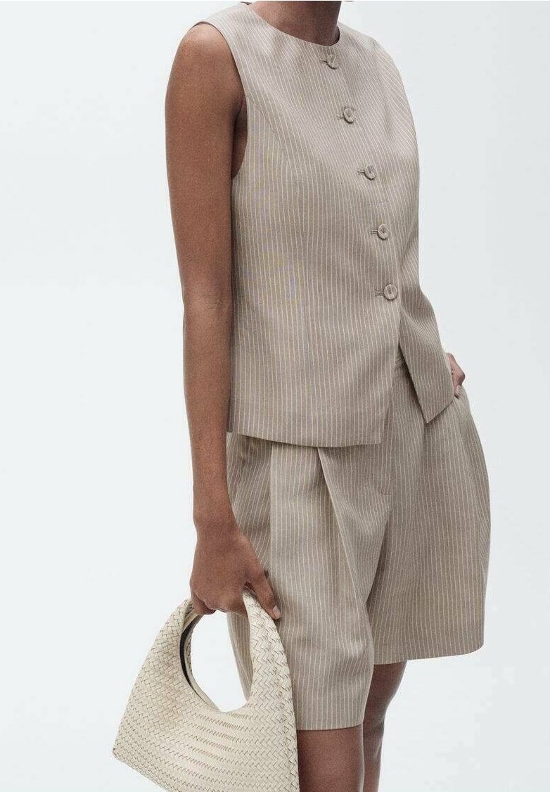 Haut beige sans manches à fines rayures avec boutons, assorti à un short mi-long, tenant un sac à main tissé crème sur un fond uni.