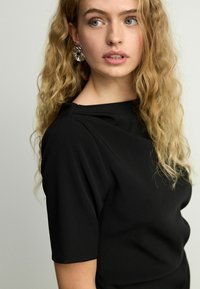 Haut noir à manches courtes, texture lisse et décolleté structuré. Cheveux coiffés en boucles lâches, portant de grandes boucles d'oreilles métalliques.