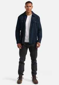 Dunkelblaues Hemd mit zwei Brusttaschen, über einem weißen T-Shirt getragen, kombiniert mit schwarzen Jeans und braunen Stiefeln.