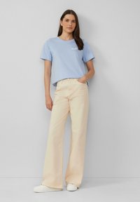 T-shirt en coton bleu clair avec le texte "HAPPY", associé à un pantalon large crème et à des baskets blanches. Design simple, coupe décontractée, texture lisse.
