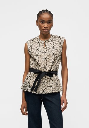 Femme portant un haut beige sans manches à motifs avec des lignes abstraites noires, une ceinture noire à la taille et un jean bleu foncé sur un fond uni.