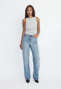 Frau mit langen, glatten Haaren, die ein ärmelloses graues Top, hellblaue hoch taillierte Jeans und schwarze spitze Schuhe trägt, steht vor einem weißen Hintergrund.