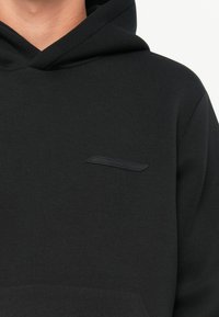 Gros plan sur une personne portant un sweat à capuche noir uni avec un petit écusson rectangulaire noir sur la poitrine gauche.