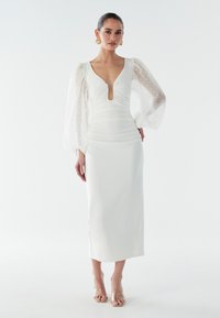 Vit figursydd klänning med djup V-ringning och rynkad front. Genomskinliga puffärmar med prickar. Mjukt, elegant tyg. Transparanta klackar i blockform.