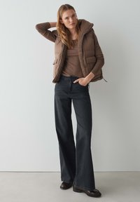 Braune gepolsterte Weste mit einer mit Fell gefütterten Kapuze, getragen über einem braunen Langarmoberteil und dunklen Schlagjeans. Geschlossene Stiefel runden das Outfit ab.