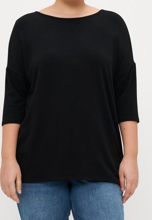 Person wearing a loose black boat neck top with elbow-length sleeves and blue denim jeans, standing against a plain background.