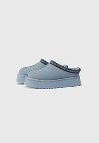 Heledad sinipäid ja platvormiga kingad tumedatest sinistest õmblustest ning UGG logo küljes, paigutatud valge tausta peale kõrvuti.