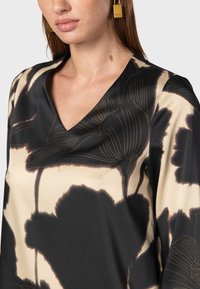 Top nero e beige con scollo a V, realizzato in un materiale liscio, decorato con grandi motivi floreali in tonalità scure. Orecchini dorati come accento.