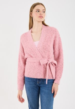 Pink knitted cardigan with long sleeves, wrap design, tie belt at the waist, and textured fabric. Worn over a light top and paired with jeans.