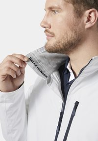 Chaqueta blanca impermeable con un cuello gris forrado de forro polar. Presenta cremalleras en color marino y la marca "HELLEY HANSEN" en el interior del cuello.