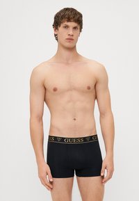 Černé boxerky vyrobené z bavlny, s zlatým logem "GUESS" na pásku, těsný střih a klasický design.