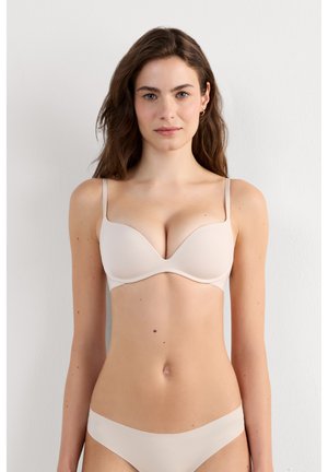 Kvinde med langt brunt hår iført matchende beige bh og trusser, står mod en plain hvid væg og vender fremad.