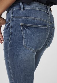 Persona vestida con jeans de mezclilla azul, con un bolsillo trasero visible, junto a parte de una mano descansando al lado, contra un fondo sencillo.
