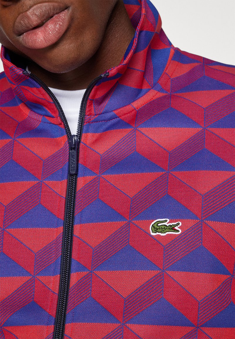 Veste zippée rouge et bleue avec un motif géométrique, fabriquée en tissu texturé, ornée d'un petit logo Lacoste vert sur le devant.