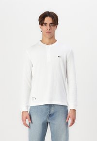 Weißes Henley-Shirt mit langen Ärmeln und drei Knöpfen, gerippte Textur, Lacoste-Logo auf der Brust, kombiniert mit hellblauen Jeans.
