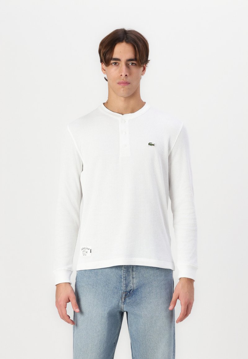 Weißes Henley-Shirt mit langen Ärmeln und drei Knöpfen, gerippte Textur, Lacoste-Logo auf der Brust, kombiniert mit hellblauen Jeans.