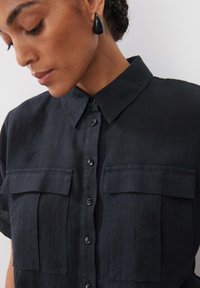 Dunkelblaues Leinenhemd mit Kragen, kurzen Ärmeln, zwei Vordertaschen und schwarzen Knöpfen. Begleitet von tropfenförmigen schwarzen Ohrringen.