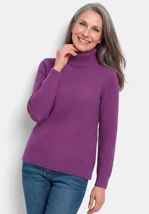 Femme souriante aux cheveux gris portant un pull à col roulé texturé violet et un jean bleu, debout devant un fond uni.