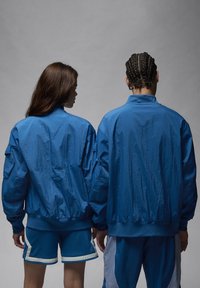 Dos personas llevan chaquetas de nailon azules con una textura arrugada. Las chaquetas tienen puños y cuellos acanalados. El fondo es gris.