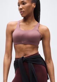 Αθλητικό σουτιέν Nike σε απαλό μωβ χρώμα, με ράντες στους ώμους και λεία υφή. Συνδυάζεται με κολάν ψηλόμεσο μπορντό και δεμένο μαύρο κάλυμμα.