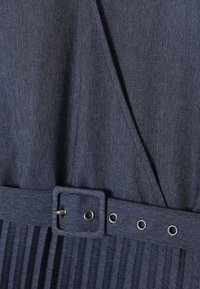 Dunkelblaues Denim-Kleid mit plissiertem Rockbereich, passendem Stoffgürtel mit Metallverschluss und Ösen, detailreiche Stickerei entlang der Kanten.
