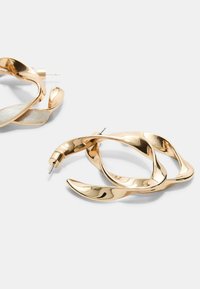 Guldtonede metal hoops med en bølget design, der har polerede overflader og nålelukninger. Subtile teksturvariationer fremhæver detaljerne.