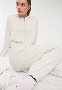 Jasnoszara bluza z kapturem na zamek błyskawiczny z przednią kieszenią oraz pasującymi dresami, o miękkiej fakturze i elastycznych mankietach, zestawione z białymi sneakersami.