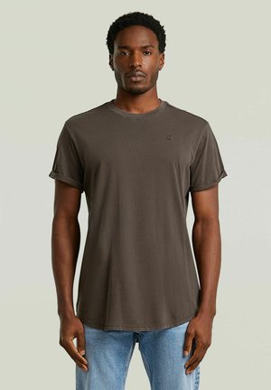 Uomo che indossa una t-shirt a maniche corte marrone scuro semplice e jeans azzurri, in piedi davanti a uno sfondo chiaro e uniforme.