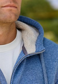 Felpa con zip di colore blu, foderata in un morbido peluche color crema. Include una camicia bianca sotto e un tessuto testurizzato, con chiusura a zip.