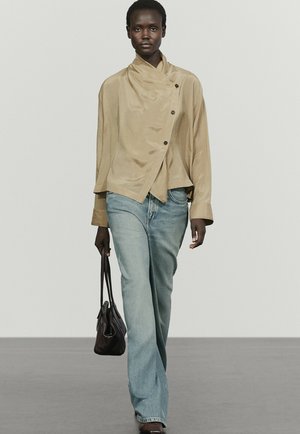 Mannequin à la peau foncée portant un chemisier beige asymétrique boutonné, un jean large bleu clair, tenant un sac à main noir, marchant sur un podium gris.