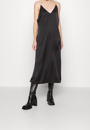 Frau trägt ein schwarzes ärmelloses Midikleid mit dünnen Trägern und schwarze kniehohe Stiefel mit Absatz vor einem schlichten hellen Hintergrund.