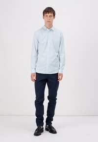 Helles blaues langärmliges Hemd mit Knopfleiste, kombiniert mit dunkelblauen Hosen und schwarzen Schuhen, was einen schlichten, maßgeschneiderten Eindruck vermittelt.