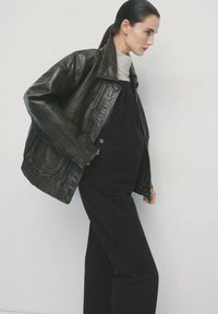 Schwarze Lederjacke mit weitem Kragen, getragen über einem grauen Pullover und schwarzen Latzhosen. Lockerer Schnitt und Seitentaschen. Glatte Textur.