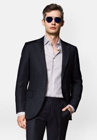 Granatowa marynarka z dwoma guzikami, zestawiona z jasną koszulą w paski. Model ma na sobie niebieskie okulary aviator. Gładki, dopasowany materiał i krój.