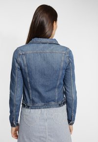 Chaqueta de mezclilla en azul claro, con un collar clásico, detalles de botones y costuras. La vista trasera muestra una silueta ajustada y mangas largas.